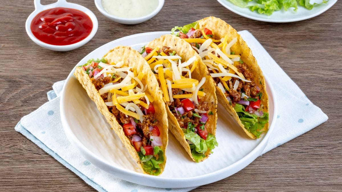 Tacoskaller fyldt med hakket oksekød, ost, hakkede løg, fintskåret tomater og agurker og majs fra dåse med salat i bunden.